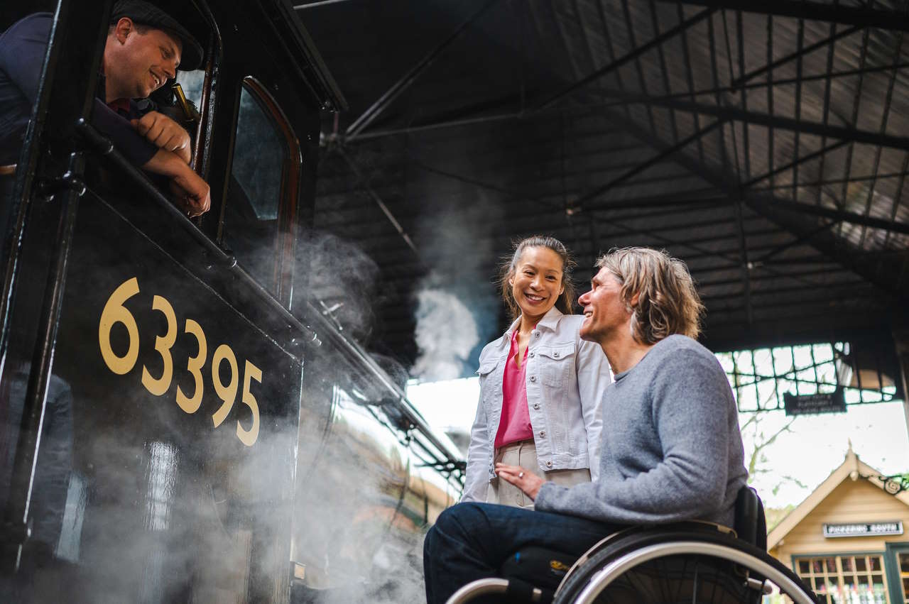 Man in a wheelchair and woman talking to the driver of a steam engine No 63395, surrounded by steam