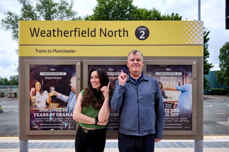 Coronation Street cast members Julia Goulding and Peter Gunn pointing at the 'Weatherfield North' tram station sign. 