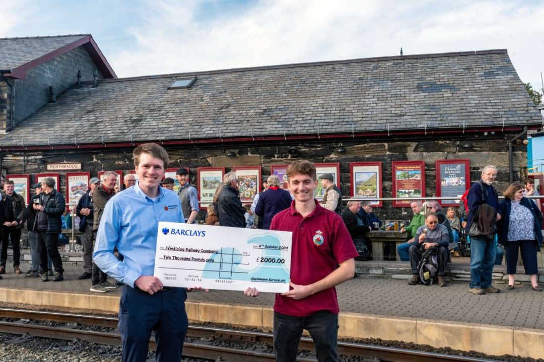 Richard Proudman, Rail Brand Manager for Bachmann Europe Plc, presents Osian Hughes, Marketing Manager for the Ffestiniog & Welsh Highland Railway, with a cheque for £2,000 in front of the former Porthmadog Harbour Station Goods Shed which now houses Spooner’s Café and Bar.